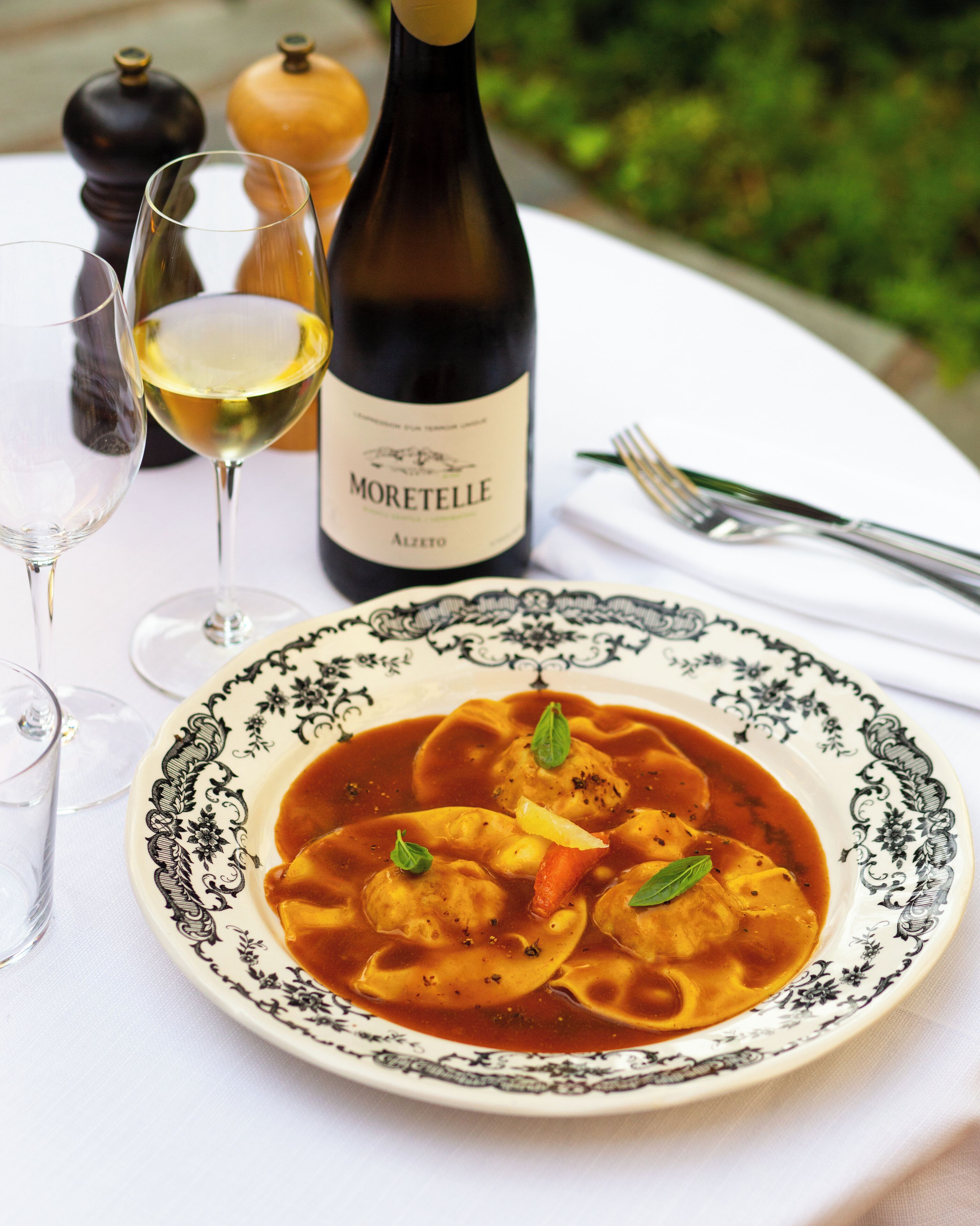 A table with a plate of ravioli in a tomato-based sauce, a bottle of Moretelle wine, and two glasses of white wine.