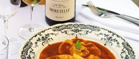 A table with a plate of ravioli in a tomato-based sauce, a bottle of Moretelle wine, and two glasses of white wine.