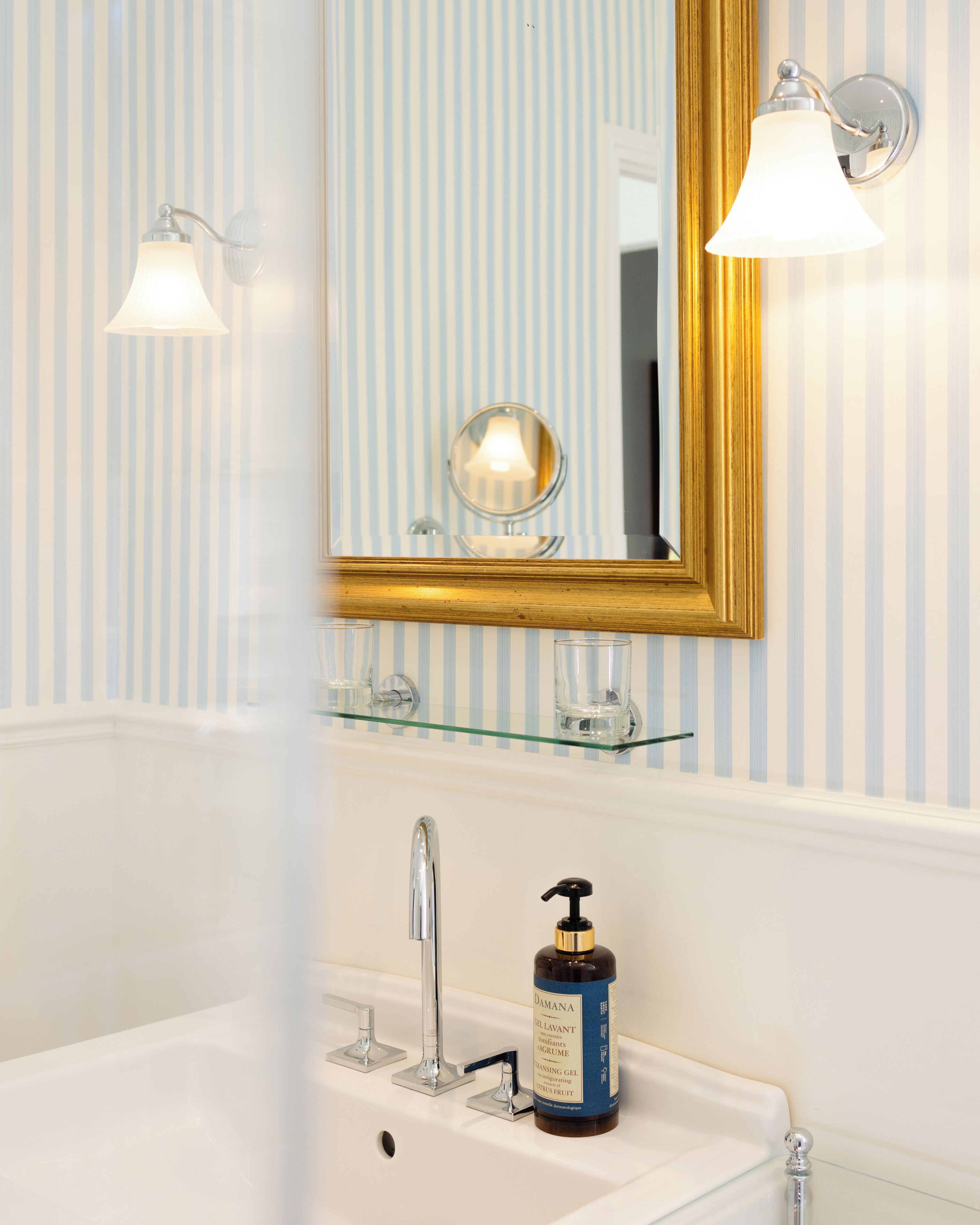 Une salle de bain avec un lavabo blanc, un miroir avec un cadre doré et une étagère en verre au-dessus du lavabo.