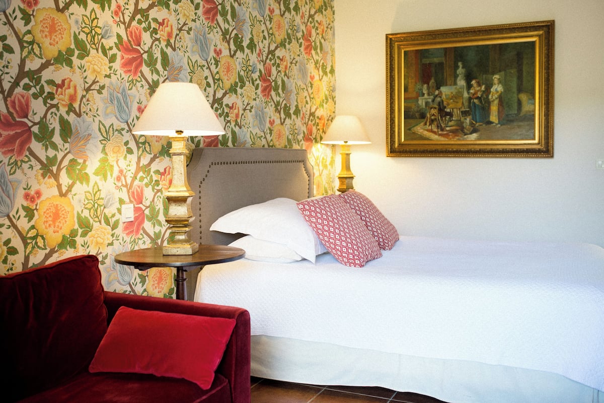 Une chambre d’hôtel avec un papier peint à motifs floraux, un lit recouvert de draps blancs, deux lampes de chevet, une chaise rouge et un tableau encadré accroché au mur.
