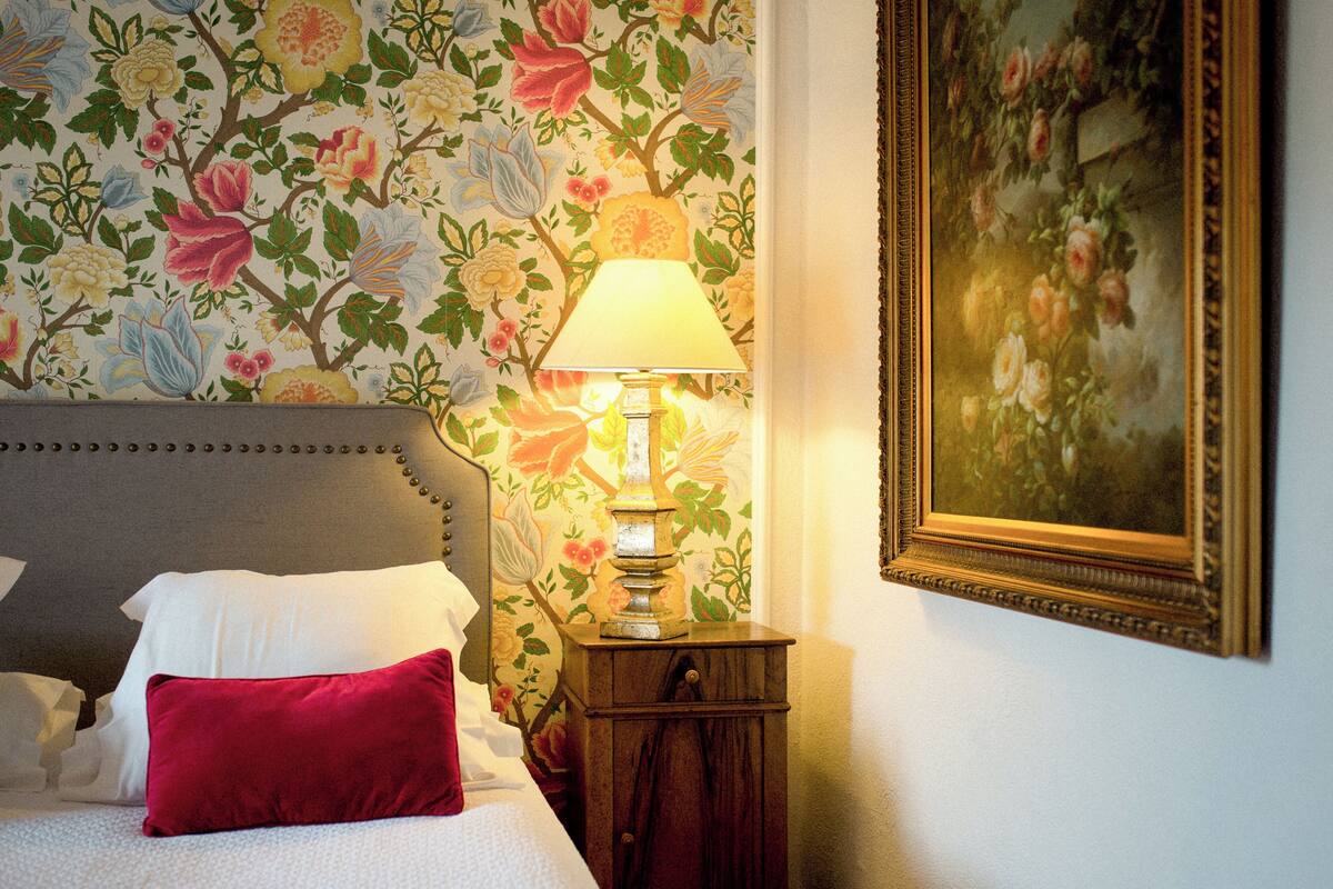 Un lit bien fait, orné d’oreillers blancs et rouges, un papier peint à motifs floraux, une lampe et un tableau encadré accroché au mur.
