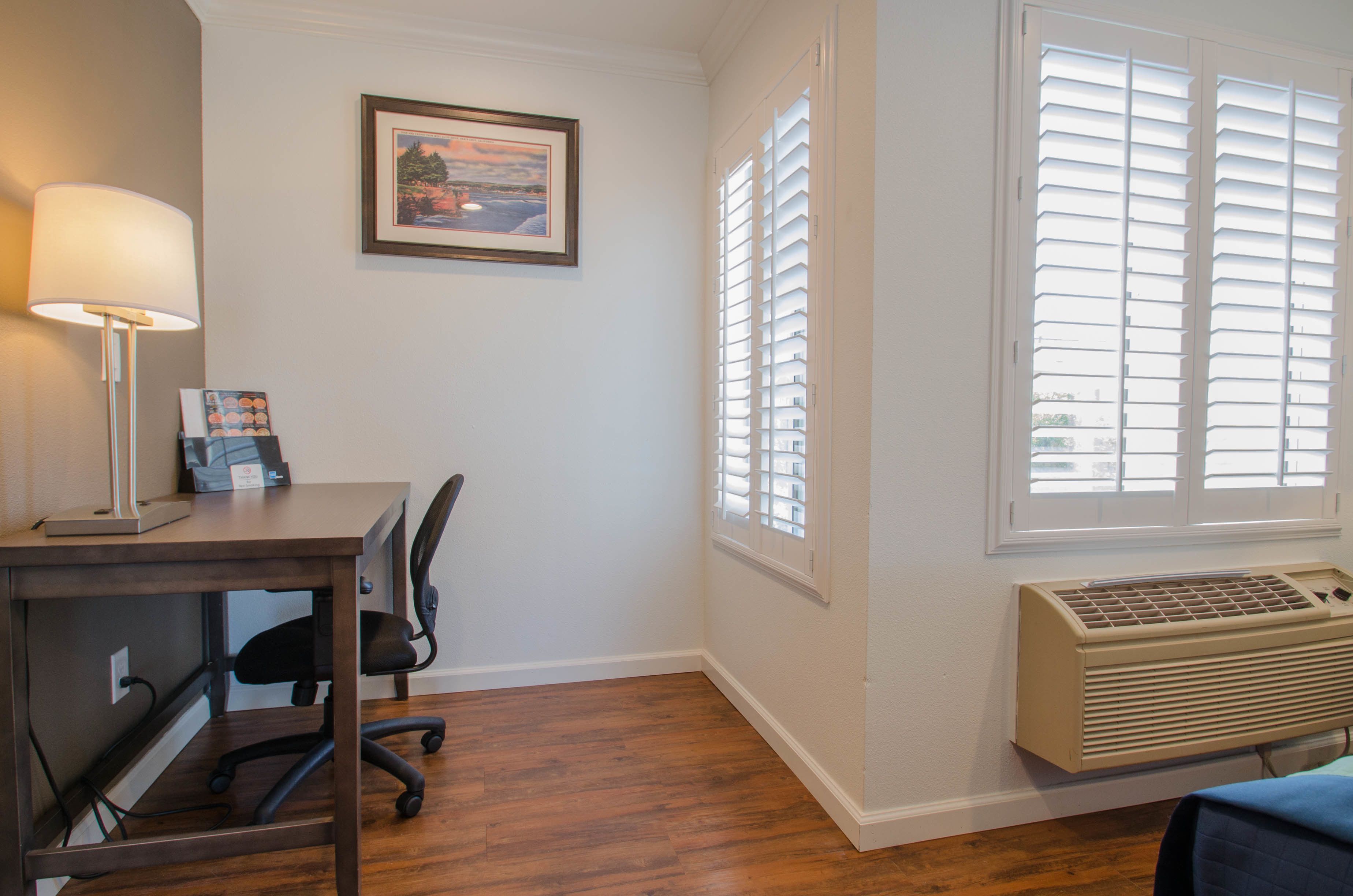 A room with a desk, chair, lamp, framed picture, and an air conditioning unit.