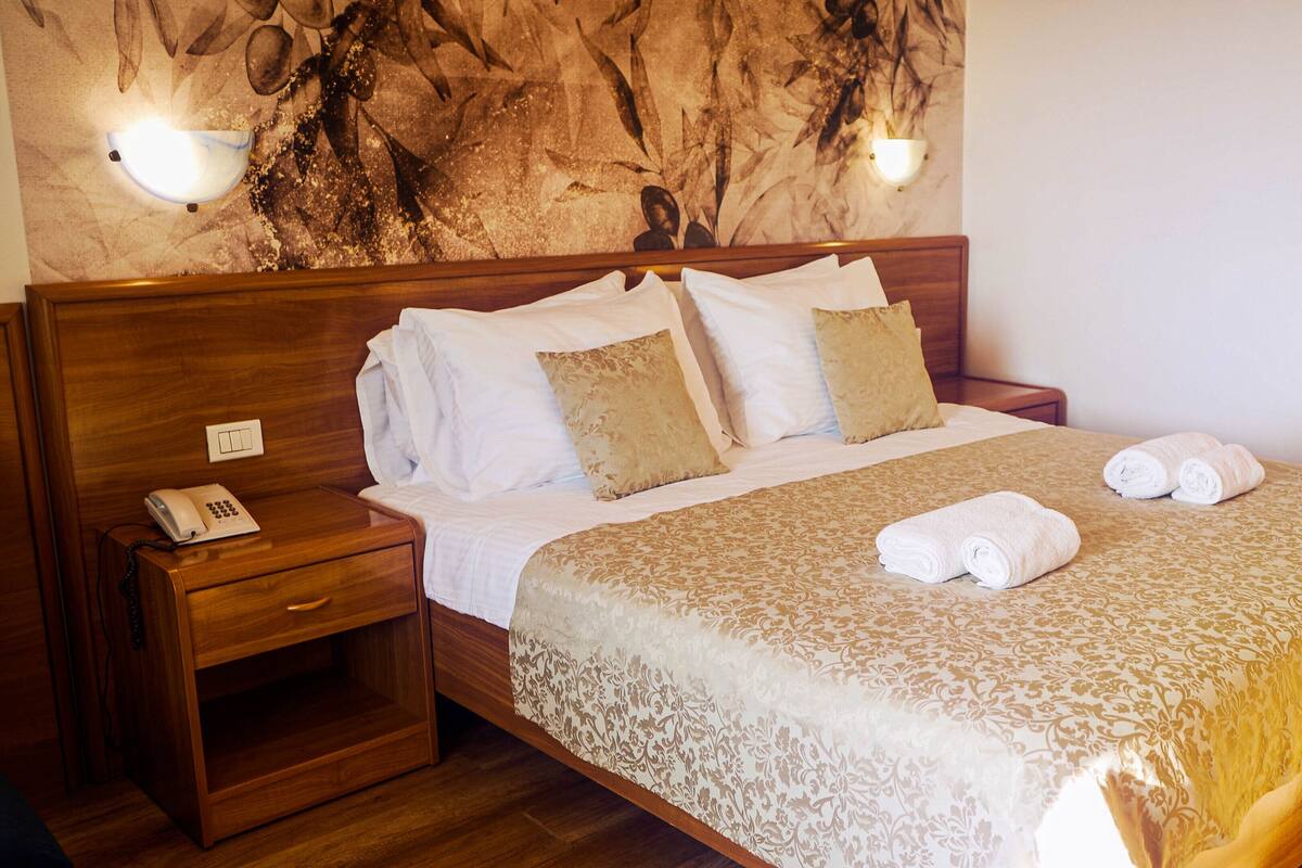 A hotel room with a wooden bed, white linens, and a patterned bedspread. There is a bedside table with a phone and a wall-mounted light fixture.
