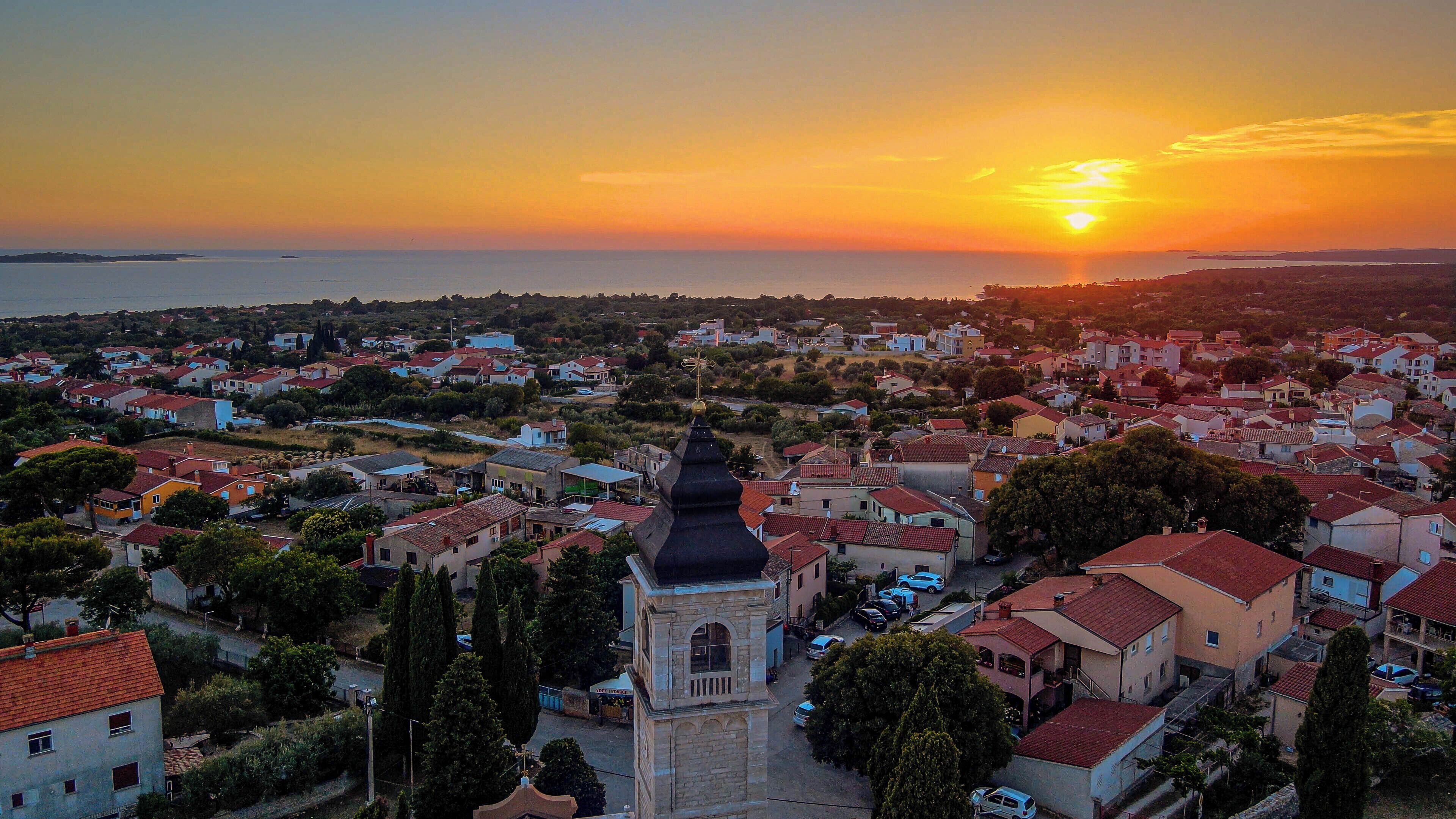 Un pueblo costero con una imponente torre de iglesia, edificios residenciales y vistas al mar al atardecer.