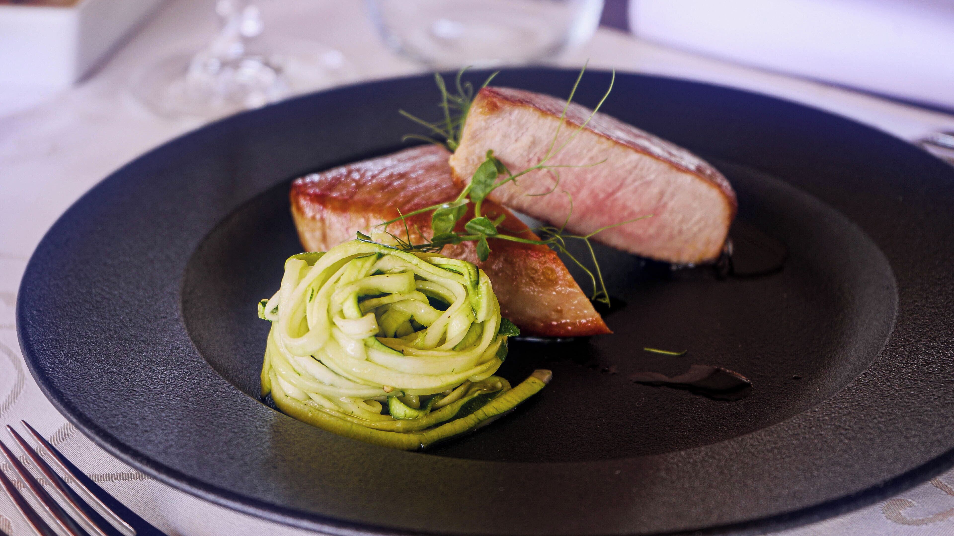A plate with a slice of meat, a spiral zucchini garnish, and a sprig of herb.