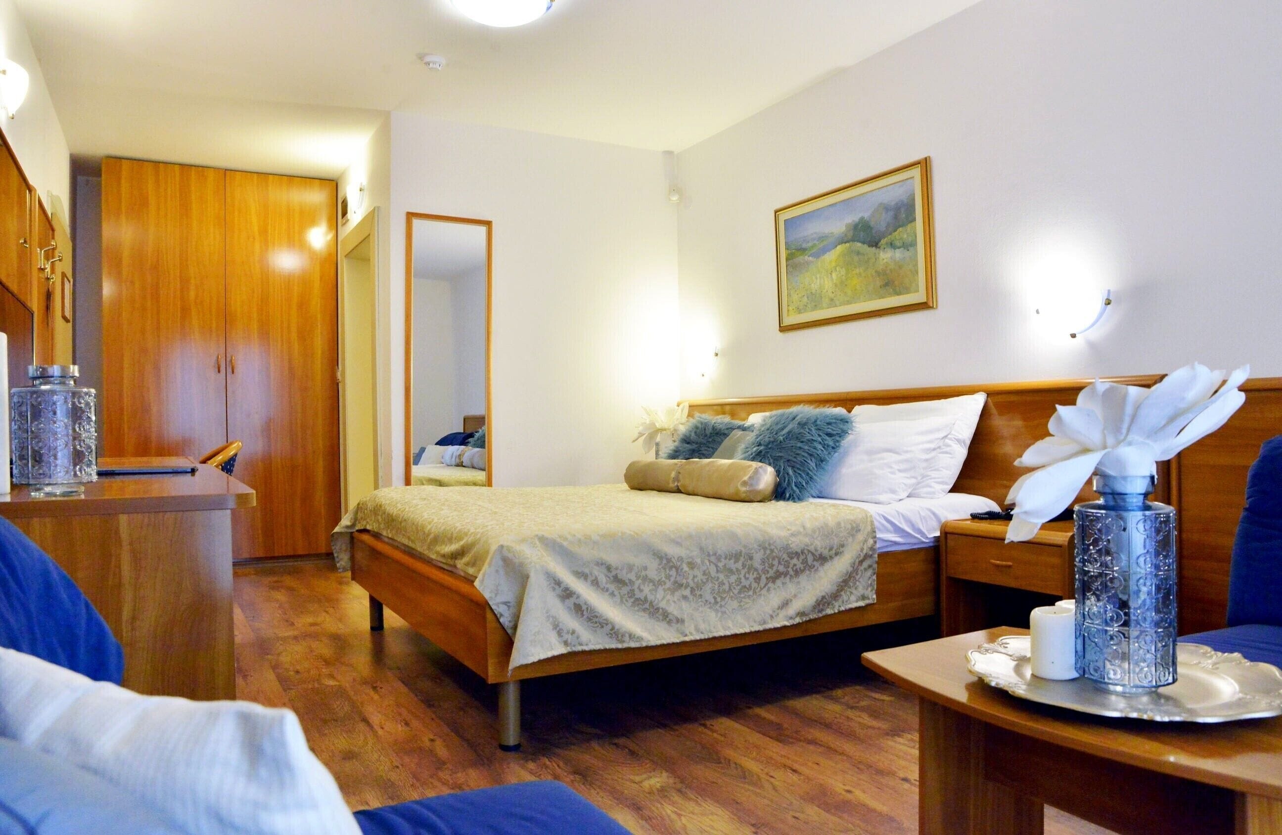 A hotel room with a wooden bed, a desk, a chair, a bedside table with a vase, and a painting on the wall.