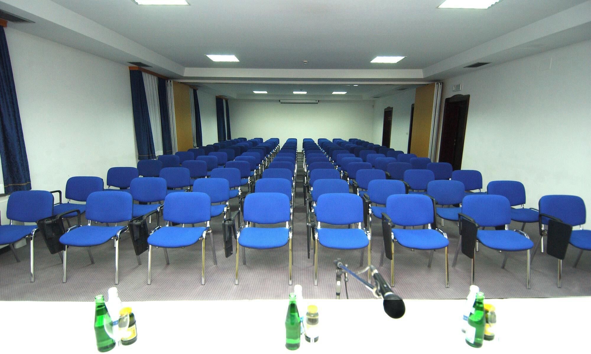 Une salle de conférence avec des rangées de chaises bleues, un pupitre et des microphones.