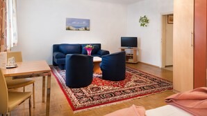 A living room with a sofa, ottomans, a table with a vase, a TV, and a red patterned rug. - Pension Dr. Geissler (Vienna)