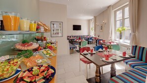 Een ontbijtbuffet met verschillende soorten fruit en drankjes, een zithoek met kleurrijke stoelen en een bank, en een aan de muur bevestigde televisie.