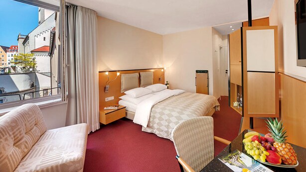 A hotel room with a bed, a chair, a table with fruit, a window with curtains, and a view of buildings outside. - Hotel am Rathaus (Augsburg)