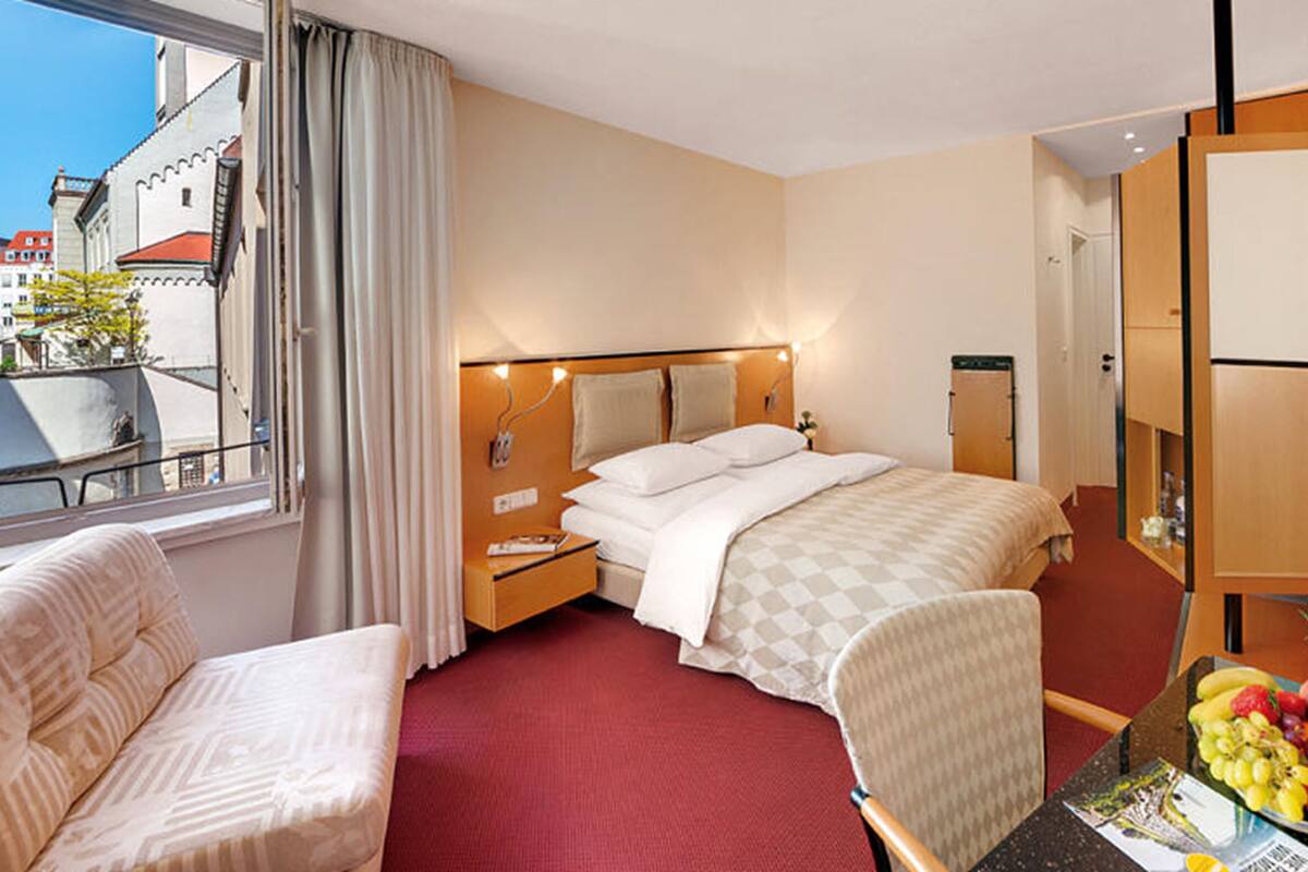 A hotel room with a bed, a chair, a table with fruit, a window with curtains, and a view of buildings outside.