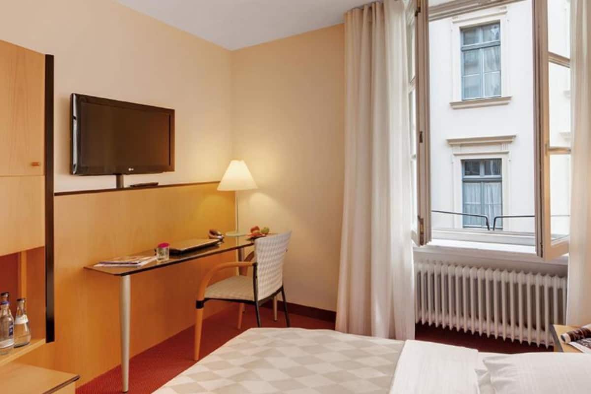 A hotel room with a bed, a desk with a chair, a television, and a window with curtains.