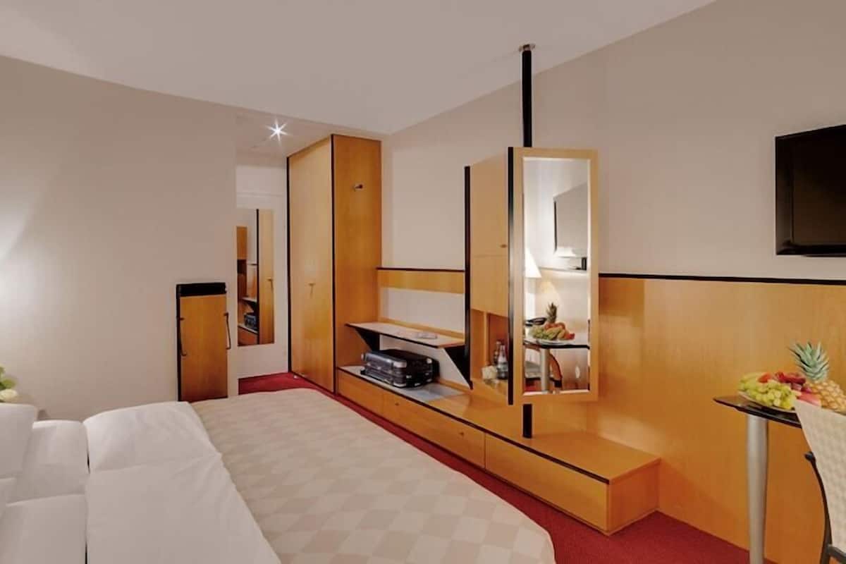 A hotel room with a bed, a desk with a mirror, a TV, and a small table with fruit.