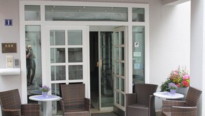 Een hotelingang met een glazen deur, rieten stoelen, een klein tafeltje en een bloempot.
