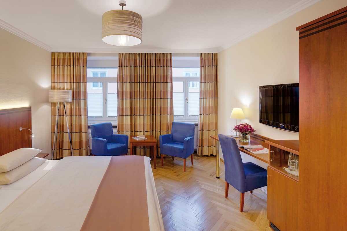 Habitación de hotel con cama, dos sillas azules, escritorio, lámpara, televisor y cortinas.