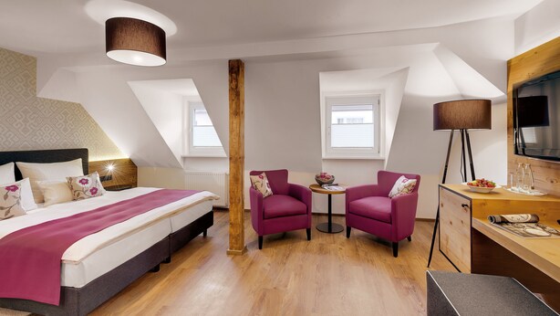 A hotel room with a bed, two armchairs, a desk, a TV, and a large window.