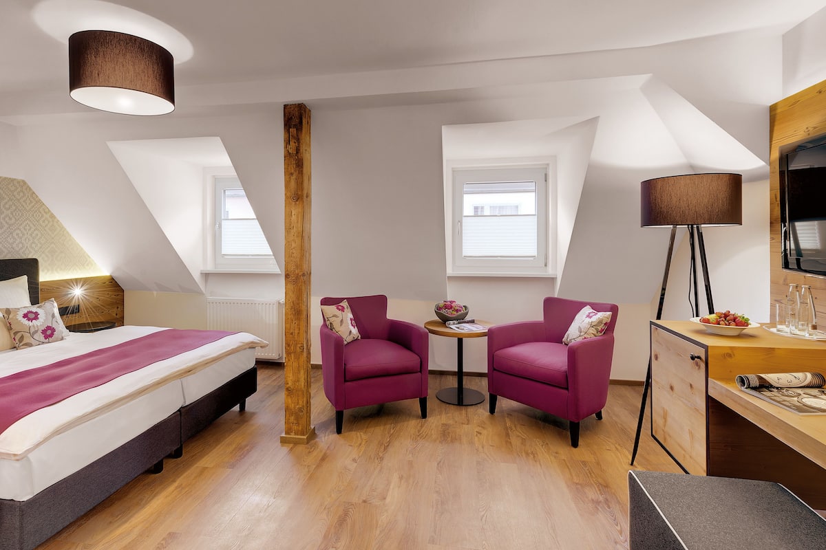 Habitación de hotel con una cama, dos sillones, un escritorio, un televisor y un ventanal grande.