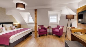 Une chambre d’hôtel avec un lit, deux fauteuils, un bureau, une télévision et une grande fenêtre.