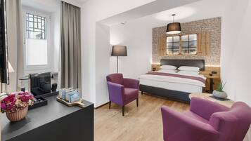 Una habitación de hotel moderna con una cama, un escritorio con cafetera, un sillón morado y una planta en maceta.