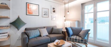 Una sala moderna con un sofá gris, una mesa de centro de madera y un sillón estampado. Hay cuadros enmarcados en la pared, un cojín verde y una lámpara.