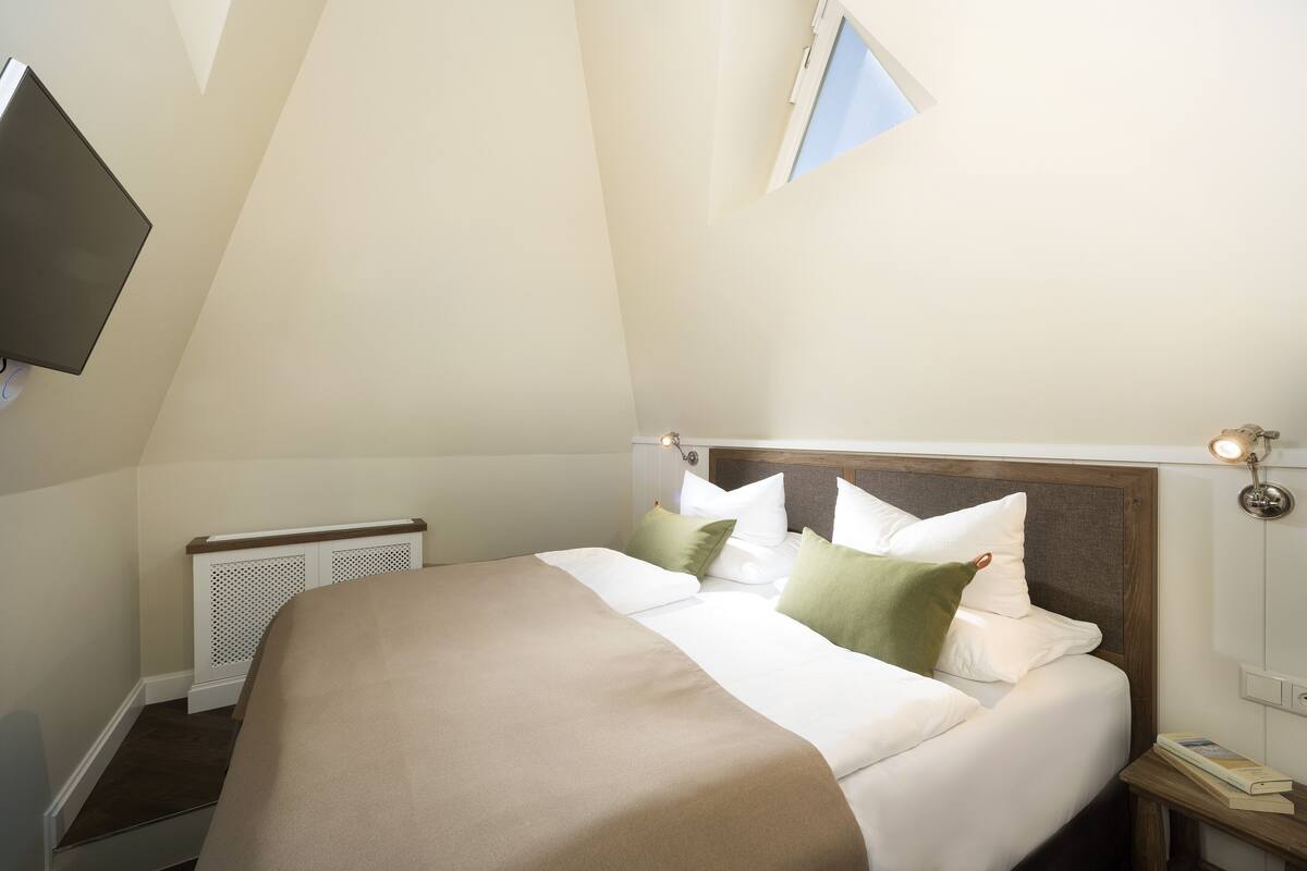 Un dormitorio con cama, mesita de noche, televisor montado en la pared, ventana y radiador.