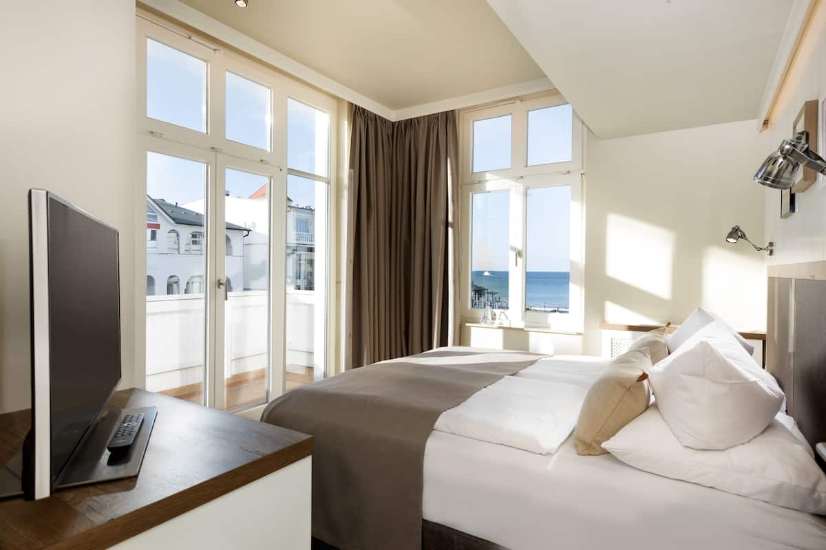 Habitación de hotel con una cama, un escritorio con una computadora portátil, un televisor y vistas al mar a través de amplios ventanales.