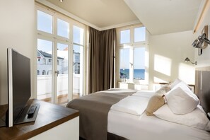 A hotel room with a bed, a desk with a laptop, a TV, and a view of the sea through large windows. - LOEV - VELA Hotel (Binz)
