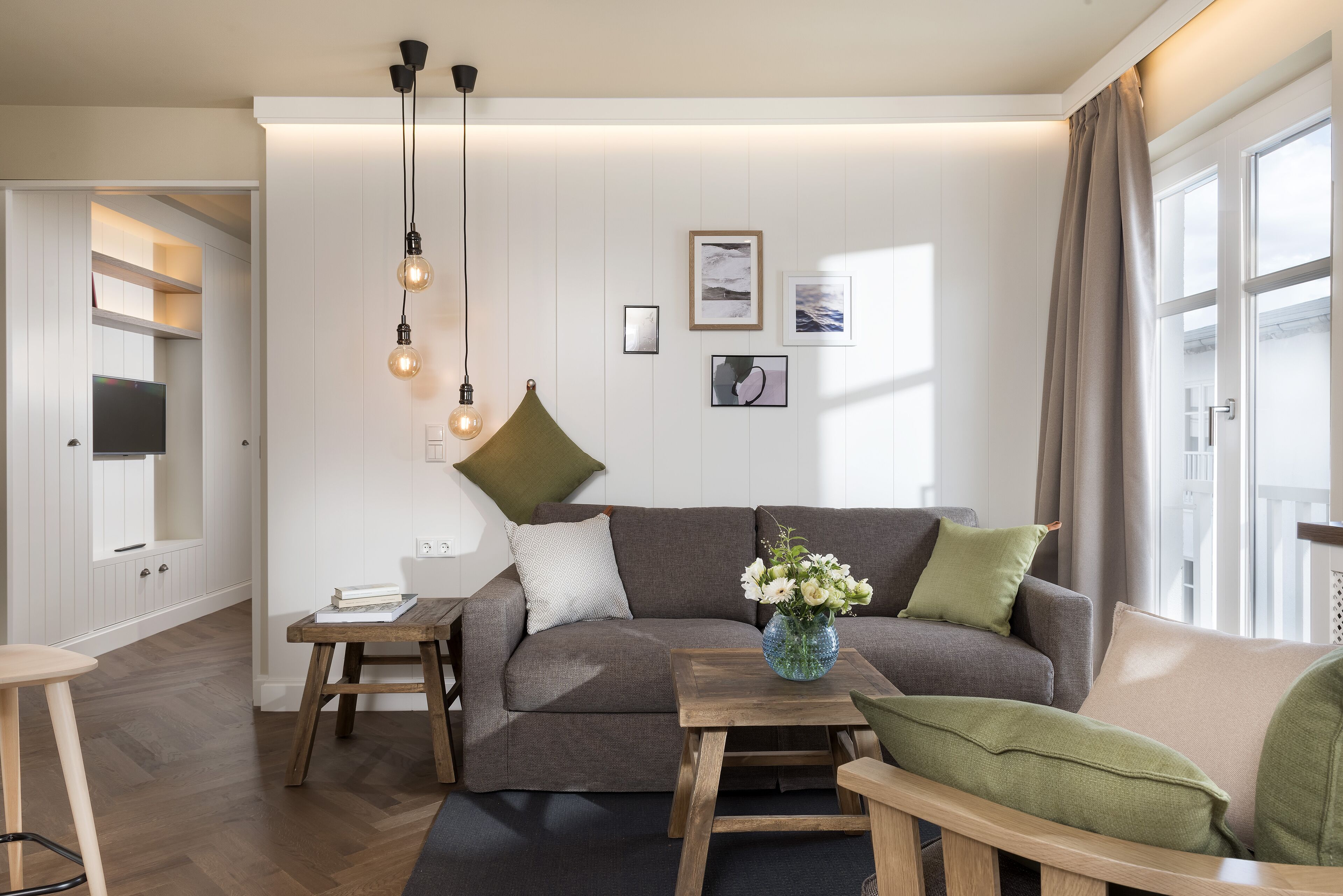Een moderne woonkamer met een bank, salontafel en hanglampen. Er is een boekenplank en een raam met gordijnen.