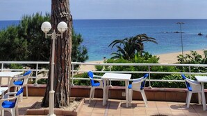 A terrace with white tables and blue chairs, overlooking a beach and the sea. - Hotel GHT Xaloc (Castell-Platja d'Aro)