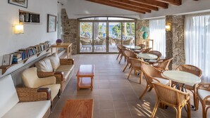 A spacious lounge area with wicker furniture, a bookshelf, and a view of the outdoors. - Hotel GHT Xaloc (Castell-Platja d'Aro)