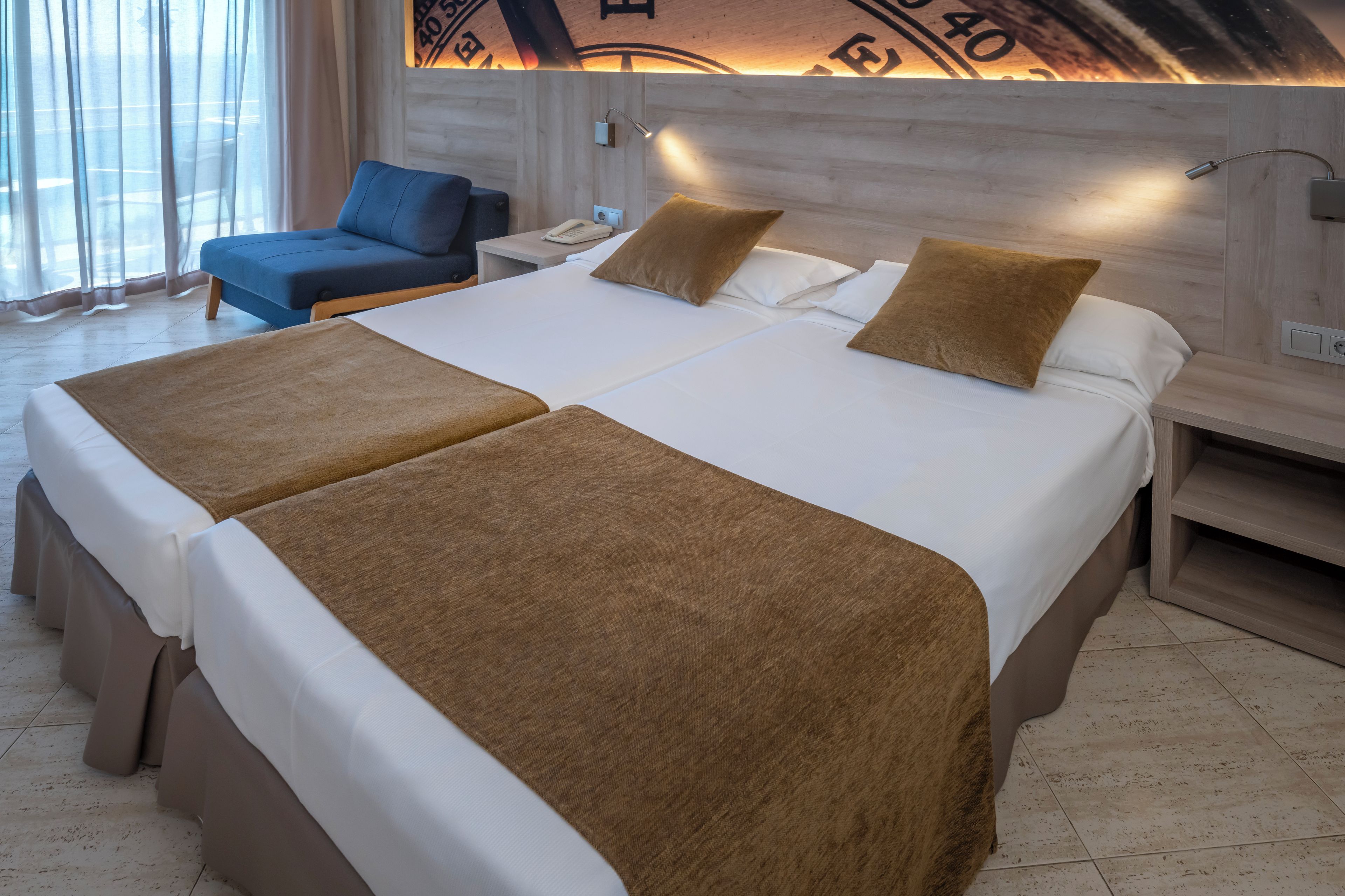 Een tweepersoonsbed met twee bruine plaids, een nachtkastje met een telefoon, en een blauwe stoel in een hotelkamer.
