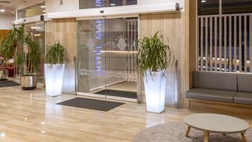 A modern hotel lobby with a reception desk, potted plants, a seating area, and a wooden floor.