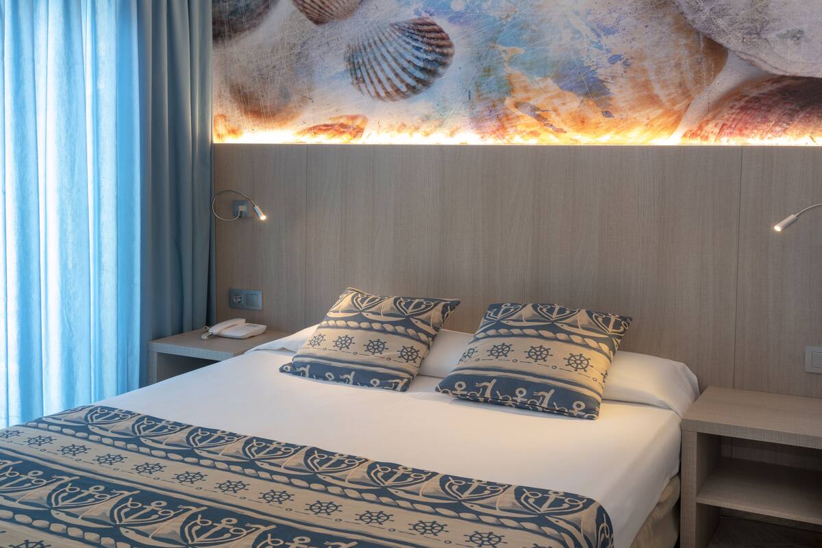 A hotel room with a bed, two pillows, a nightstand, a phone, and a wall with a shell pattern.