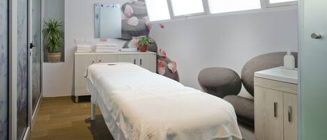 A clean, modern spa room with a massage table, a small plant, a towel rack, and a sink.