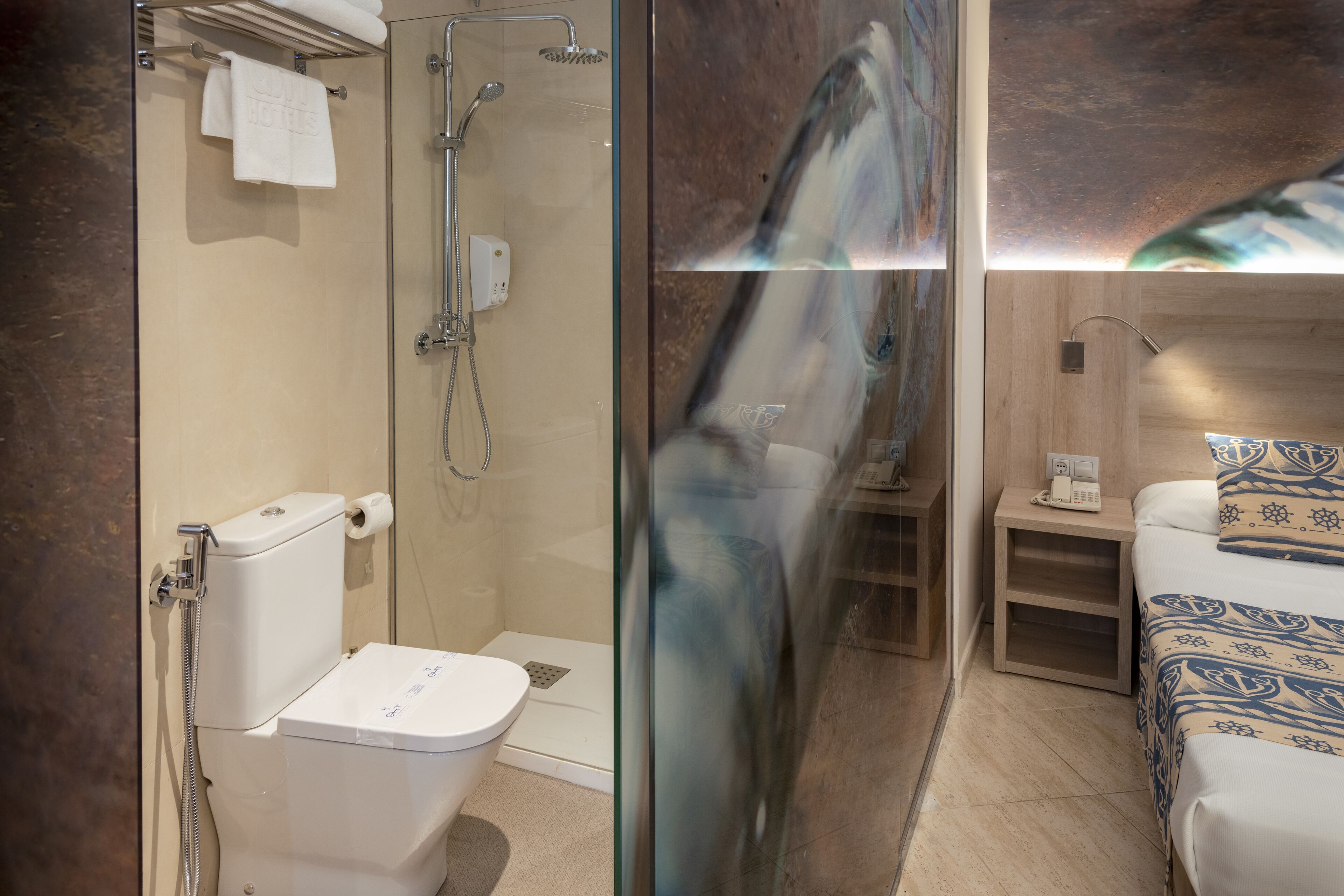 A modern bathroom with a glass shower enclosure, a toilet, and a bedside table with a lamp and a phone.