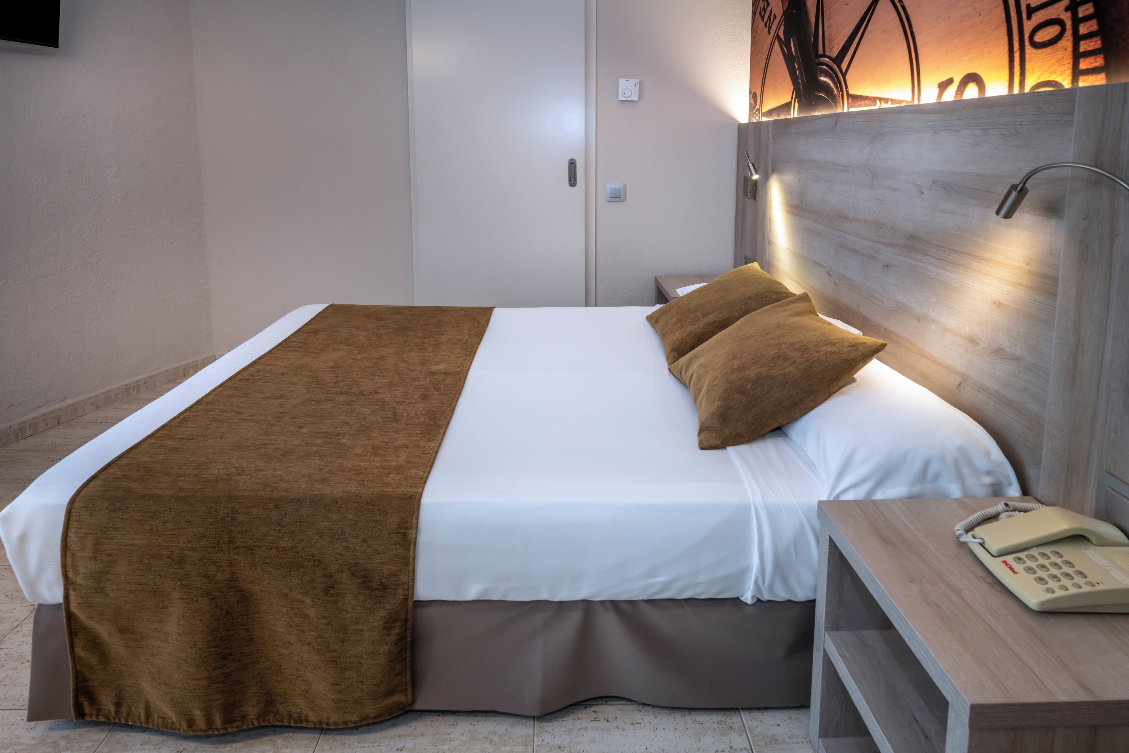 A neatly made bed with a brown blanket and pillows, a bedside telephone, and a nightstand with a lamp.