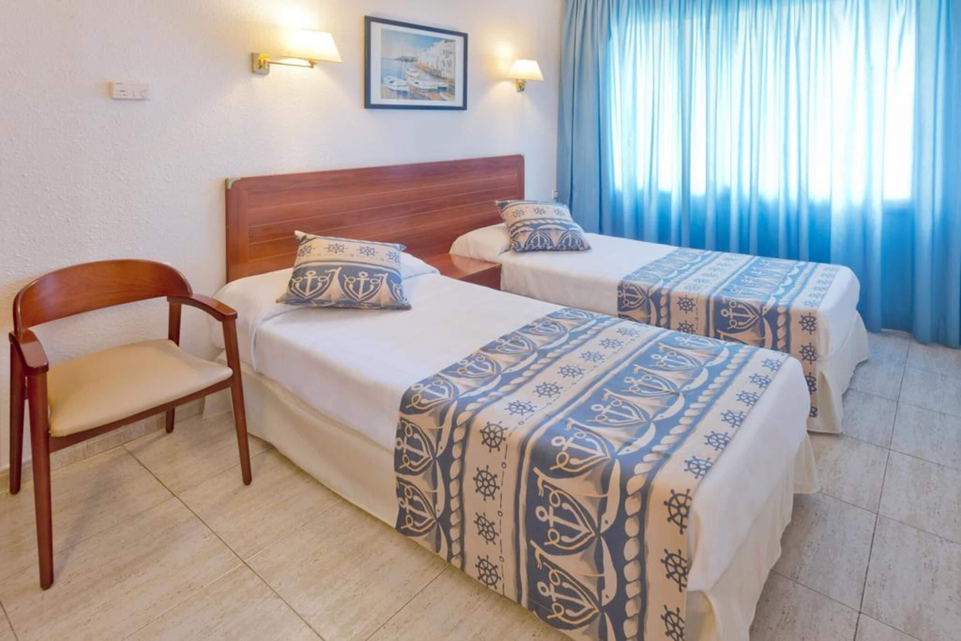 Habitación de hotel con dos camas, una silla de madera y vistas al mar a través de cortinas.