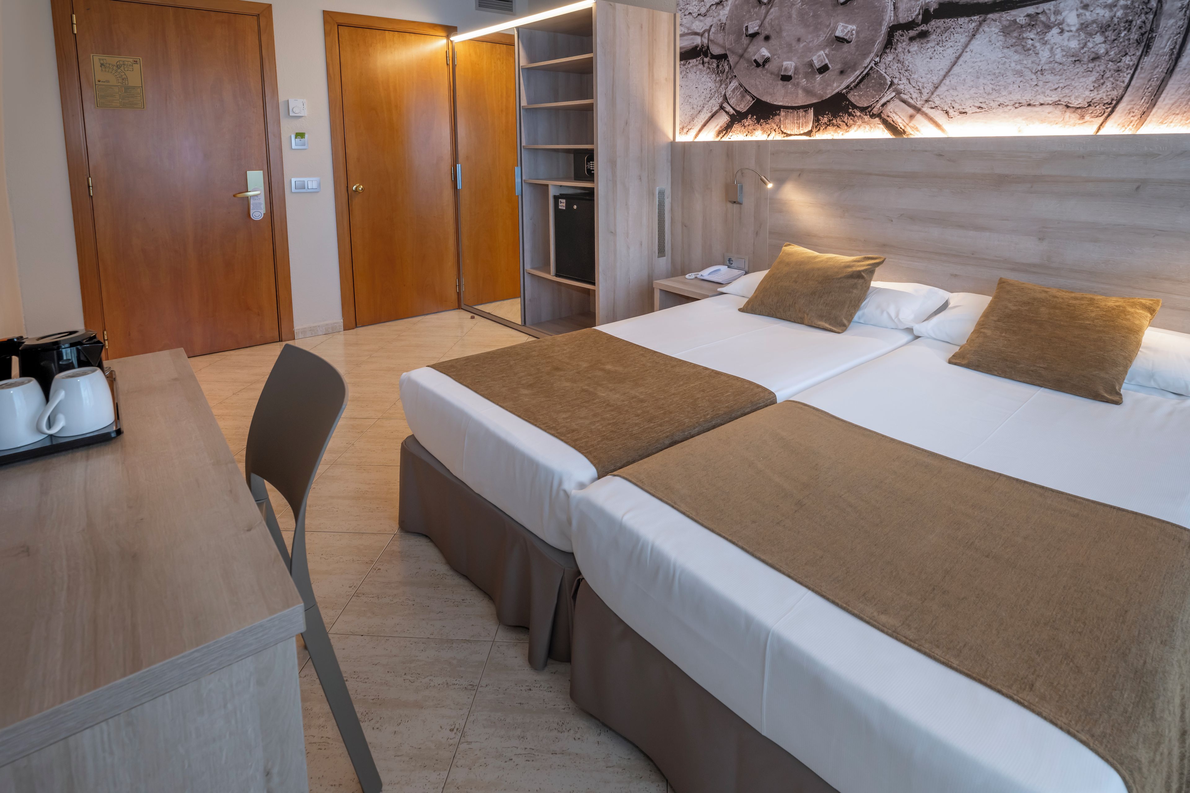 A hotel room with two beds, a desk, a chair, and a wall-mounted heater.