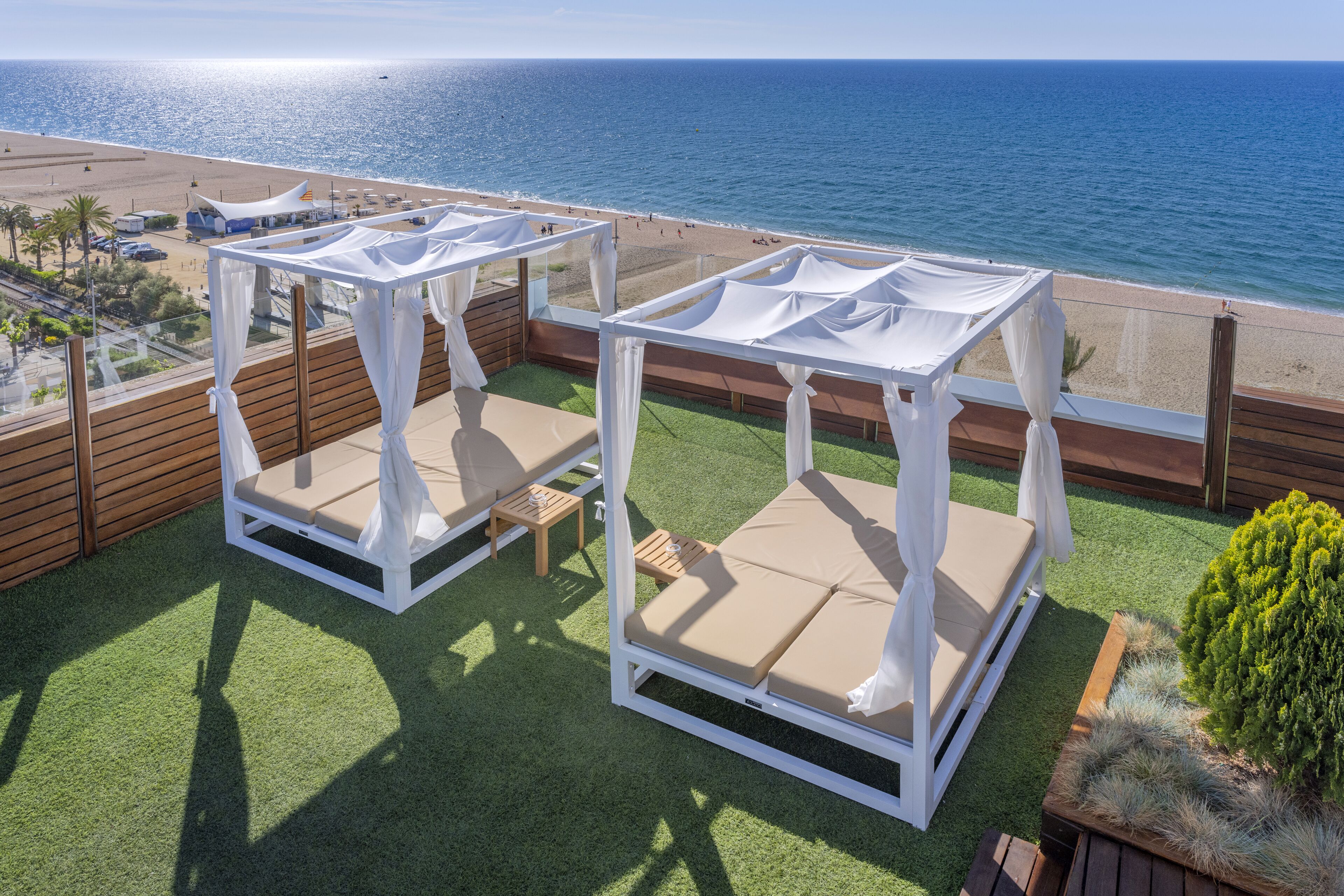 Deux espaces de détente extérieurs, aménagés avec des meubles blancs et des coussins beige, une terrasse en bois et une vue sur la plage et l’océan.
