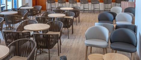 A hotel bar area with multiple seating options including round tables and chairs, a bar counter with various bottles, and a visible sign for the hotel.
