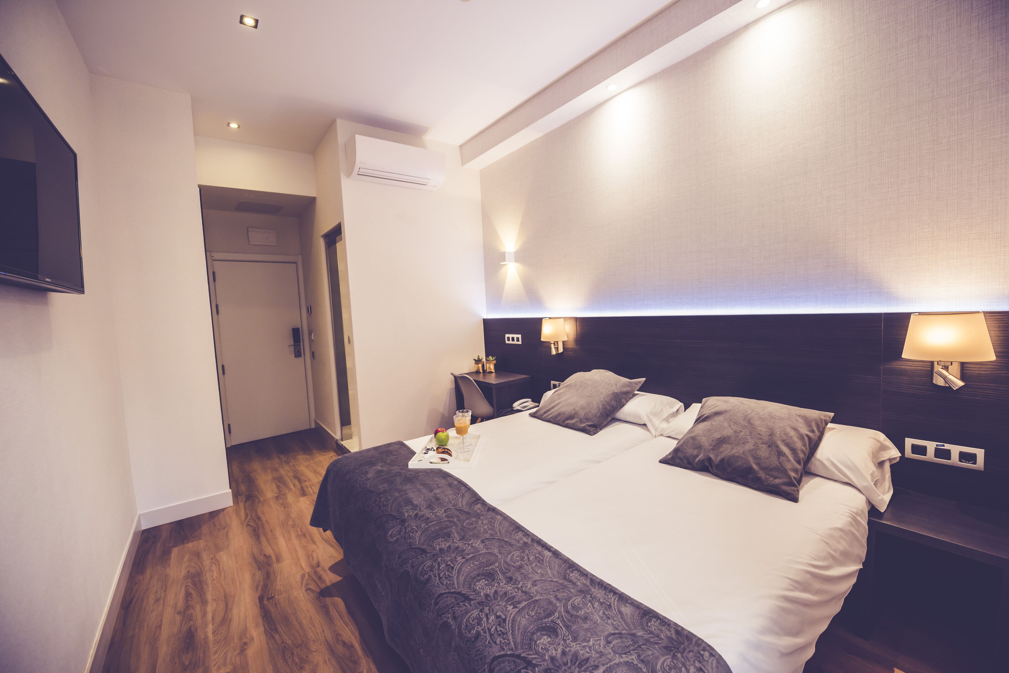 A modern hotel room with a large bed, bedside lamps, a flat-screen TV, and a wooden floor.