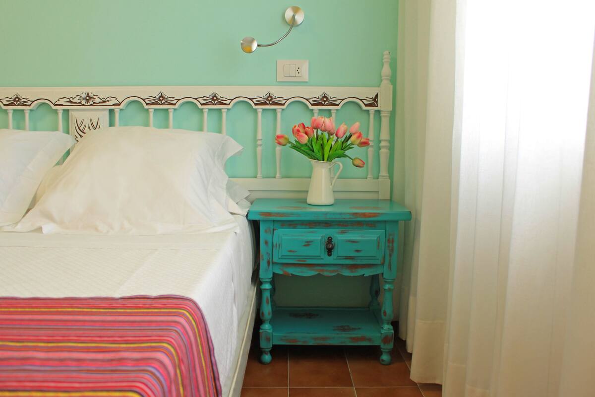 Una cama con sábanas blancas, una mesita de noche color turquesa con un florero de flores rosadas, y un interruptor de pared.