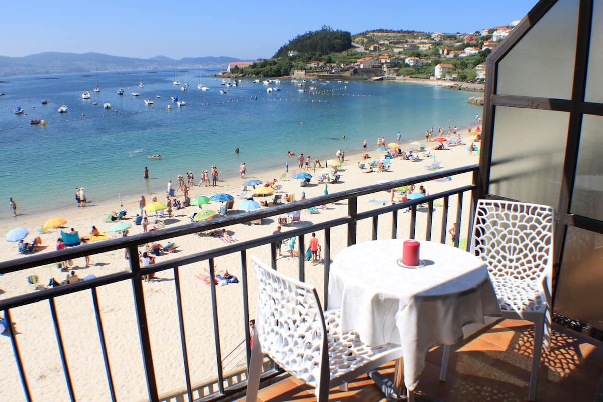 Balcón con mesa y sillas con vista a una playa con sombrillas y gente.