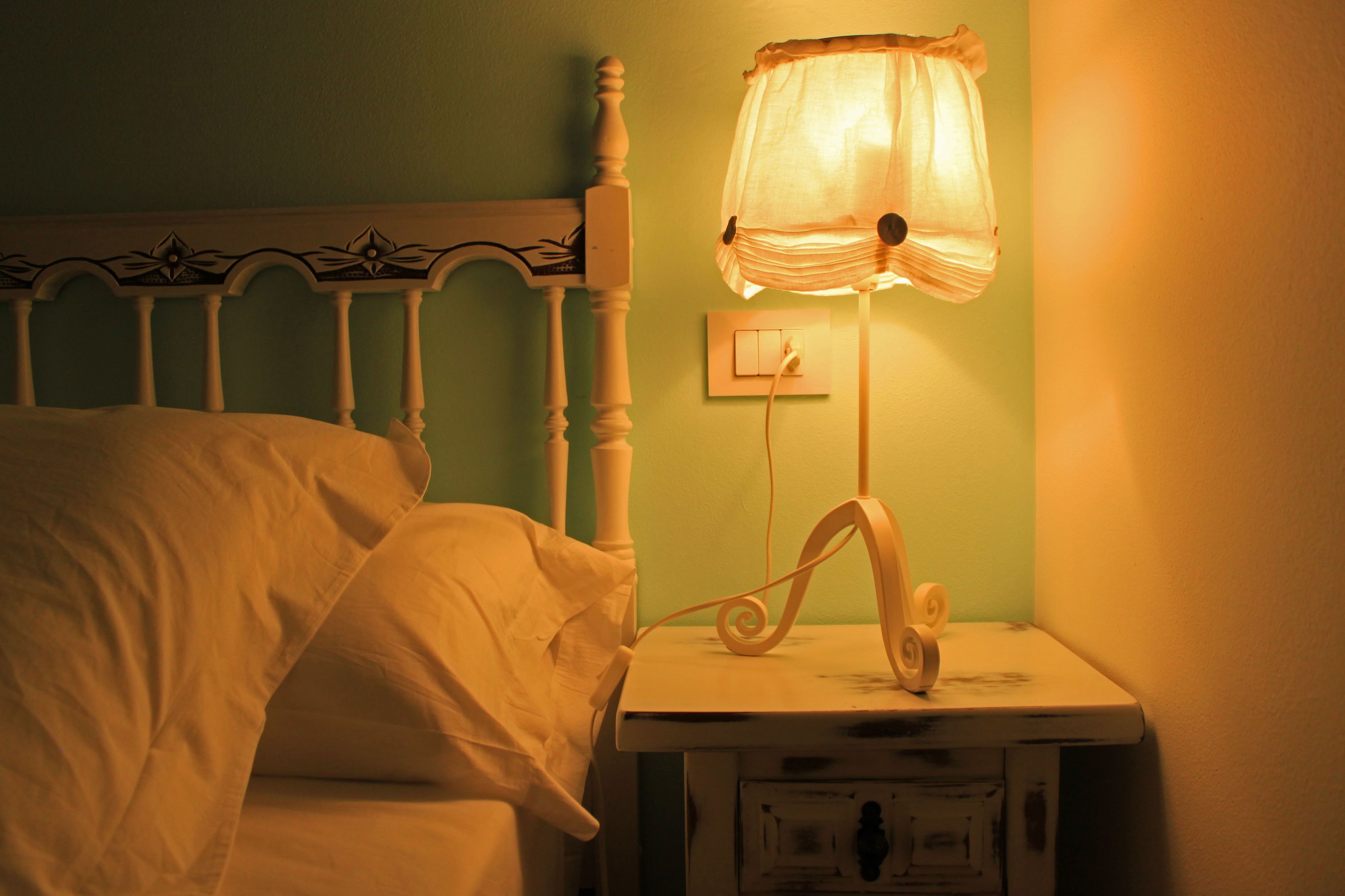 Una cama con ropa blanca, una mesita de noche blanca con una lámpara y un interruptor de pared.