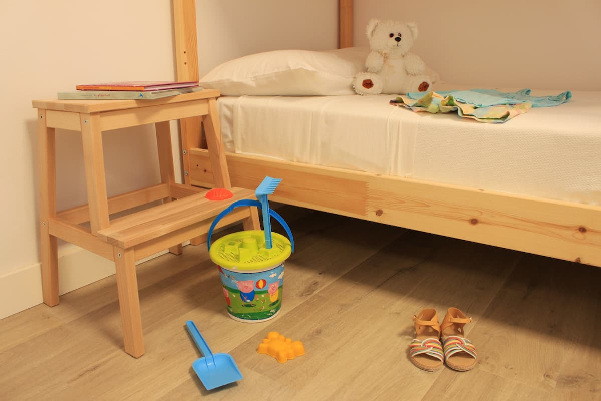 Una habitación infantil con una cama, una mesita de madera, un cubo de colores y un par de sandalias.