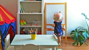 Niño dibujando en una pizarra en una habitación con una estantería, una mesa con un vaso amarillo lleno de lápices y una planta.