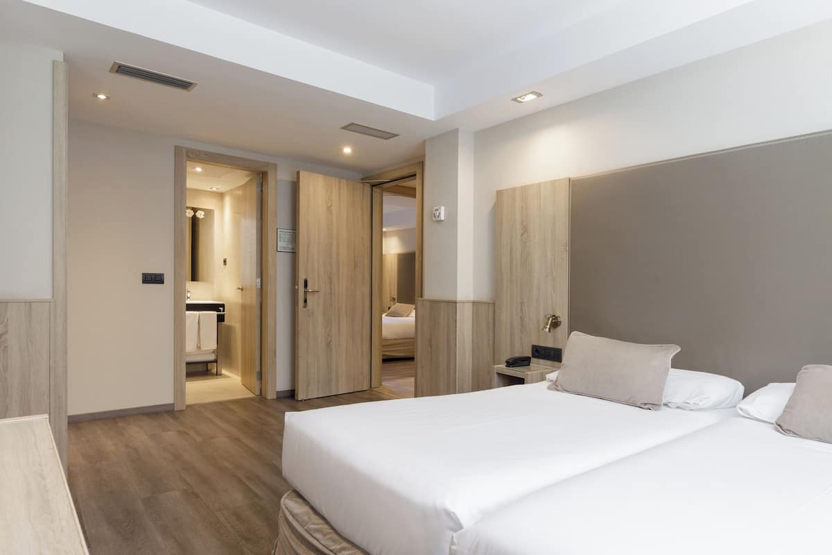 A modern hotel room with a large bed, a bedside table, a wardrobe, and a bathroom visible through an open door.