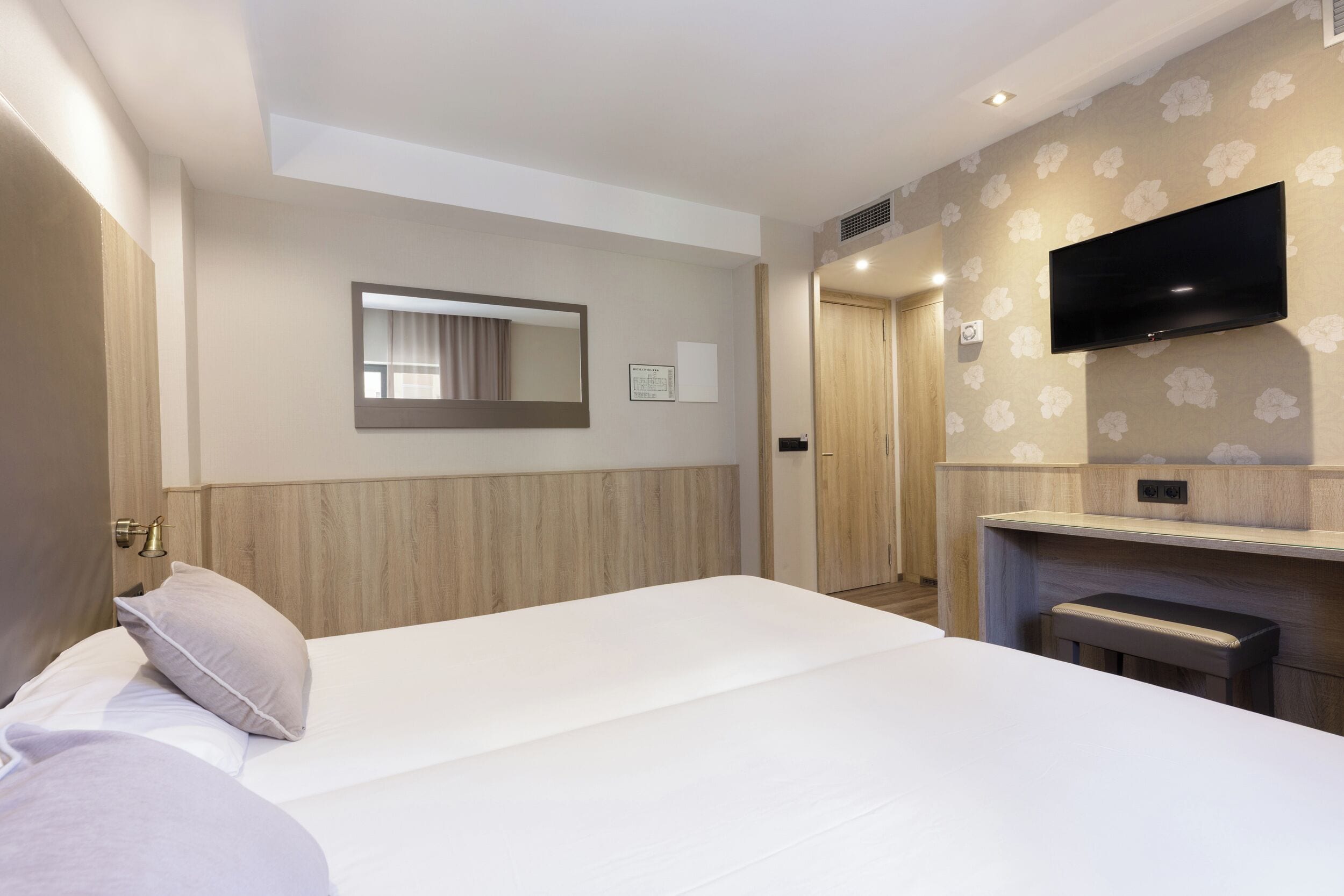 A hotel room with a large bed, a TV mounted on the wall, a wooden headboard, and a small seating area.