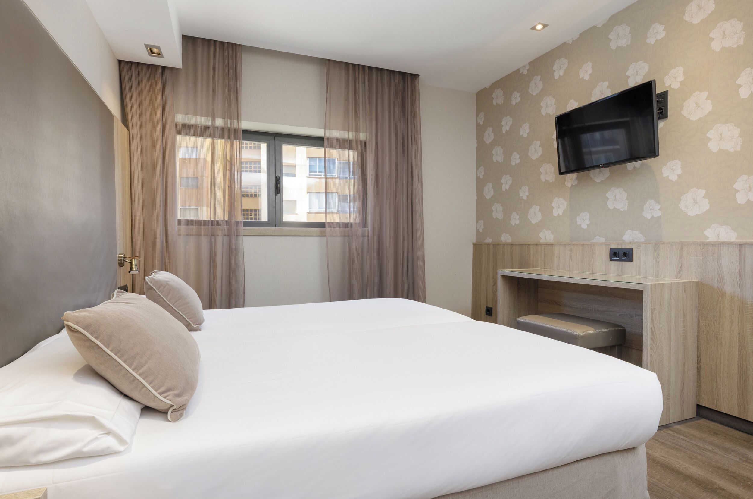 A hotel room with a bed, curtains, a TV mounted on the wall, and a window with a view of buildings.