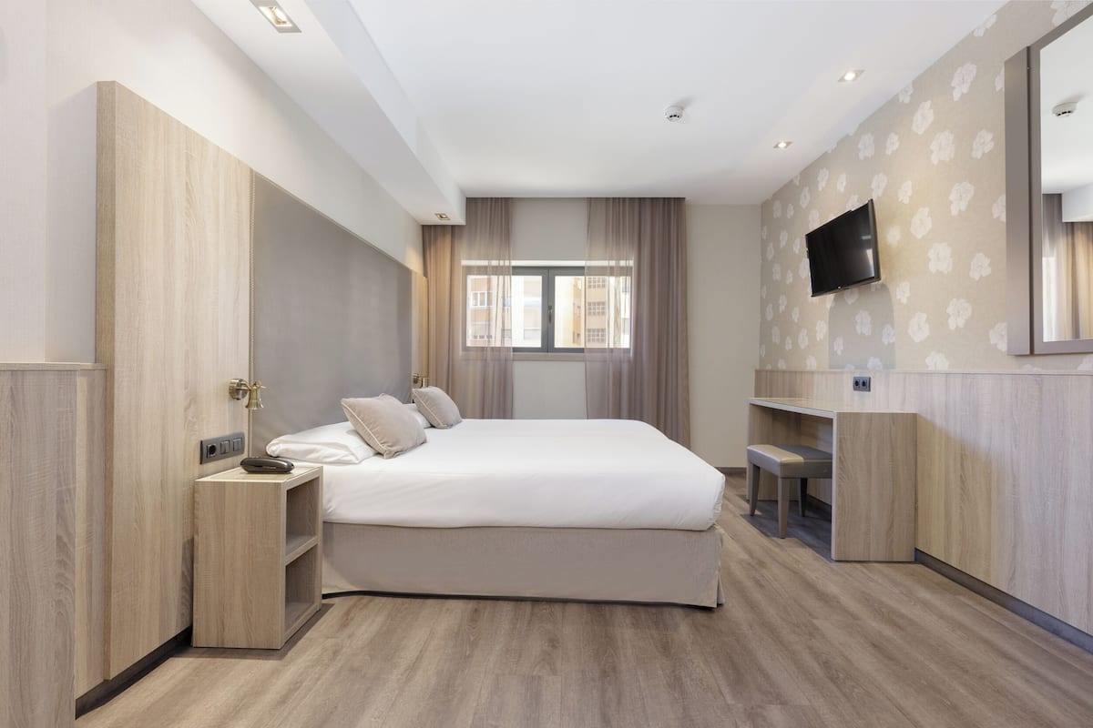 A modern bedroom with a large bed, a TV mounted on the wall, and a window with curtains.
